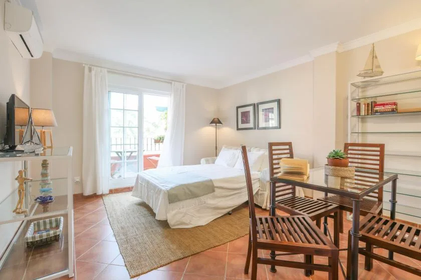 A bright bedroom with a bed, dining table, and chairs beside a glass door.