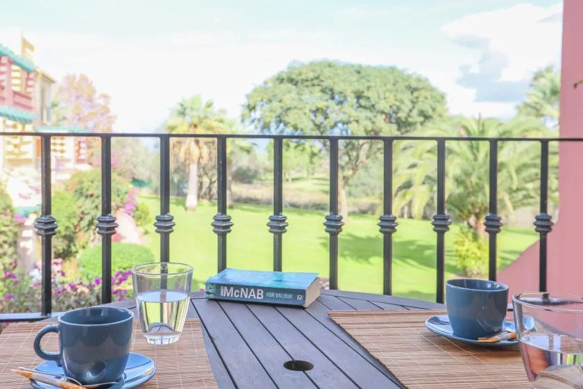 A balcony table with cups overlooks a green garden through a black railing.