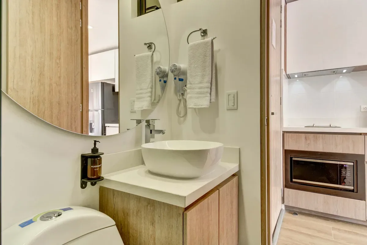 Un tocador de baño compacto tiene un lavabo blanco sobre encimera, muebles de madera, un espejo grande y toallas colgadas.
