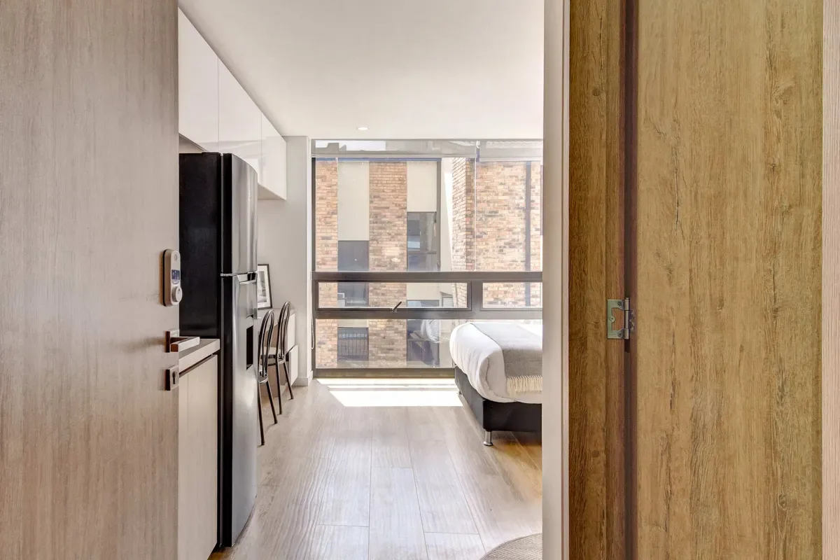 Un dormitorio compacto con una ventana grande, una cama y una pequeña mesa junto a la cocina.
