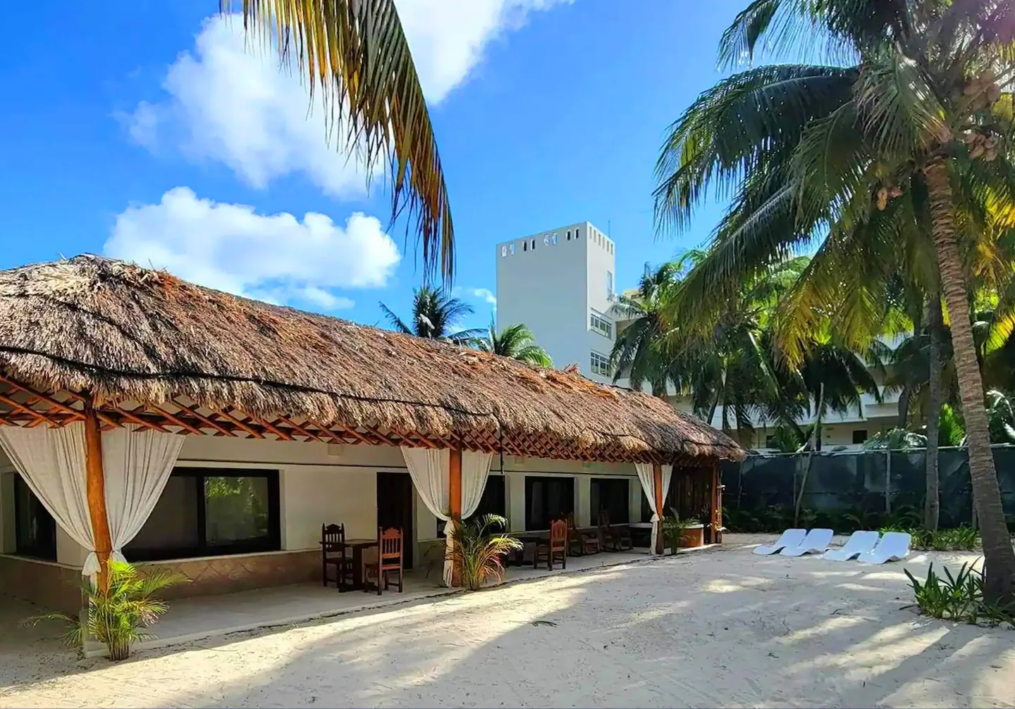 Espacio representativo del hotel: Un comedor al aire libre con techo de palma da a un patio arenoso con palmeras.