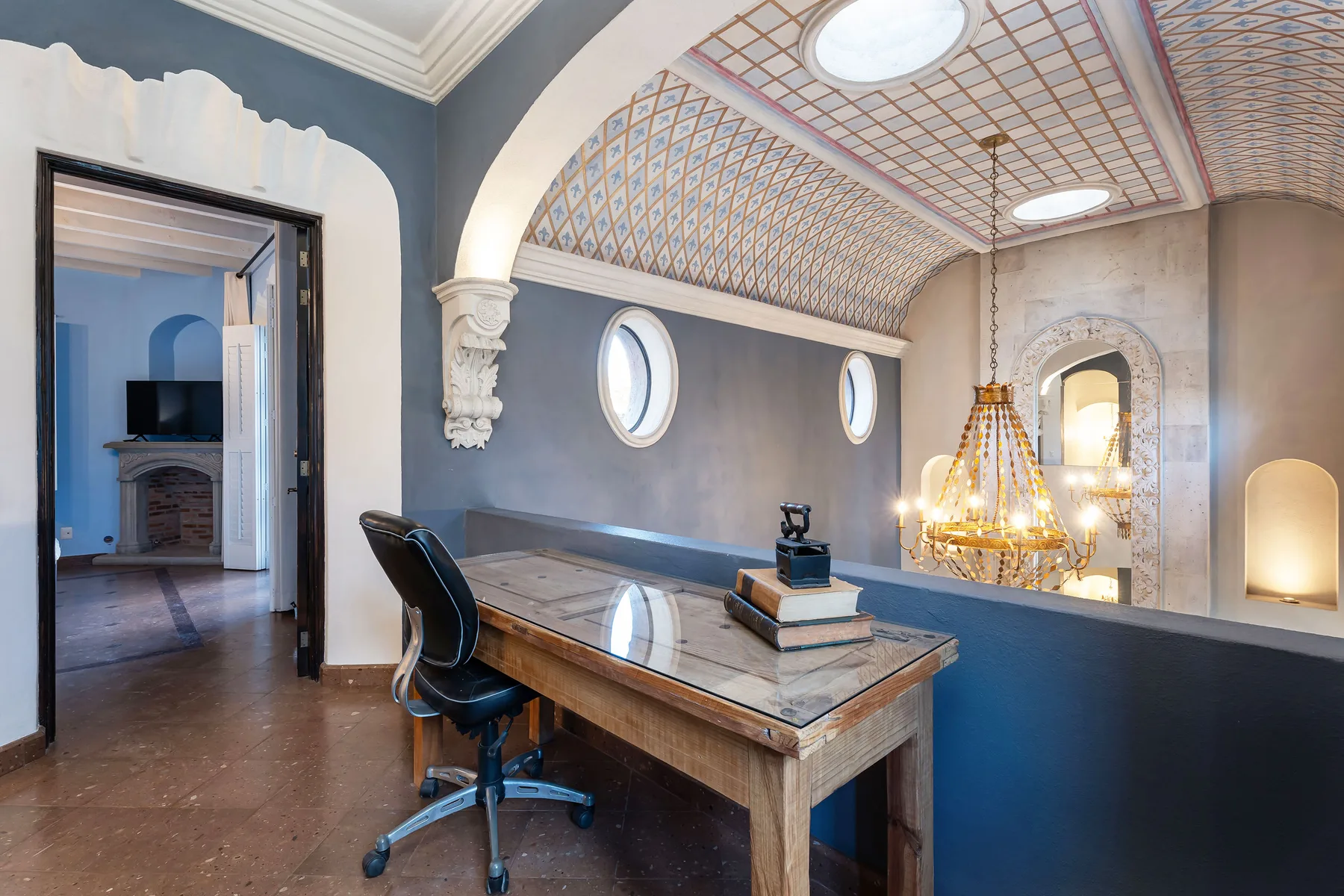 A study with a long desk and a hanging chandelier beneath a patterned ceiling.