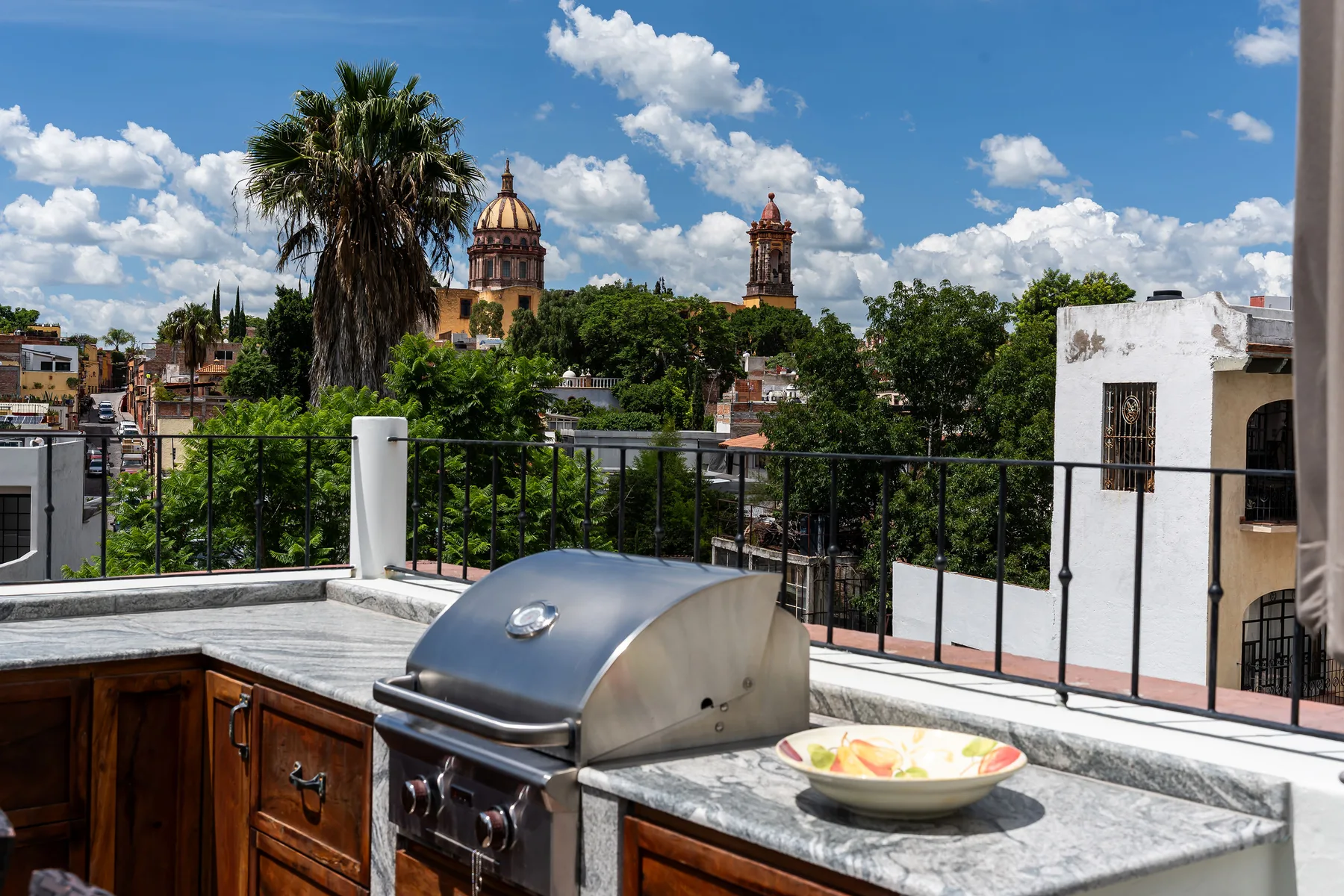 A rooftop outdoor kitchen overlooks leafy surroundings and historic towers beneath a blue sky.