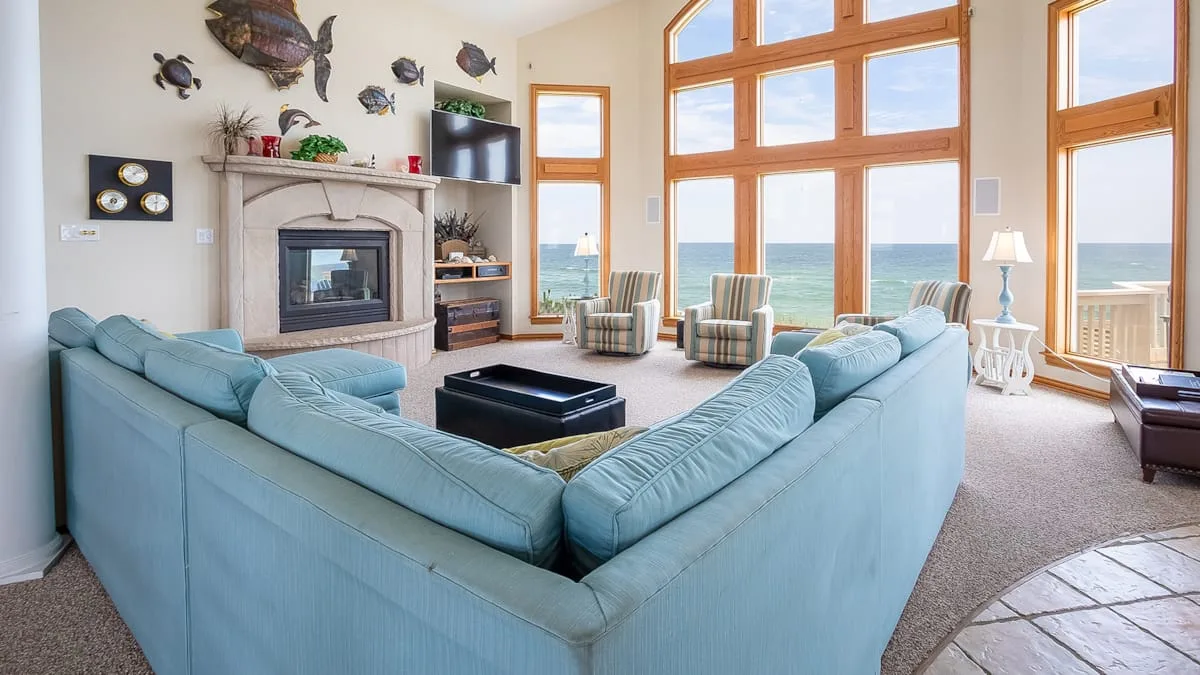 A bright living room has pale blue seating, a fireplace, and large windows overlooking the ocean.