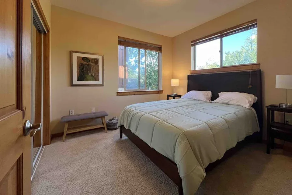 A bedroom with a bed beneath two windows and a framed picture on the wall.