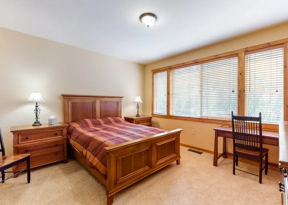 A bedroom with a wooden bed and a desk beside wide windows.