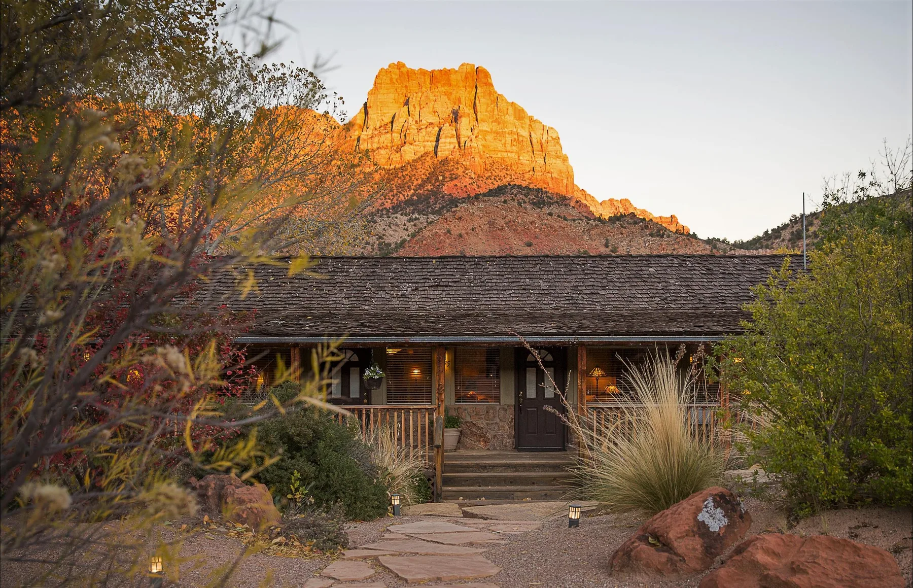 Espacio representativo del hotel: Un alojamiento rústico se encuentra bajo una formación rocosa roja iluminada por el sol, rodeado de plantas del desierto.