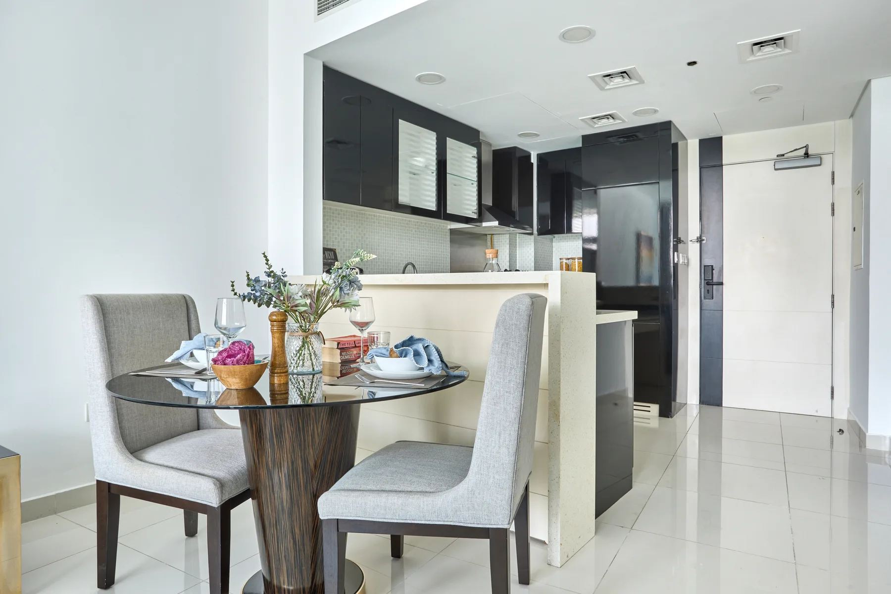Comedor con una mesa de vidrio y asientos tapizados claros junto a una cocina.