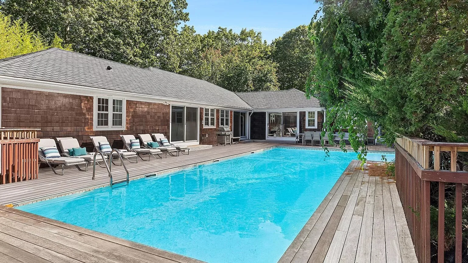 An outdoor swimming pool sits beside a wooden deck and a low building.