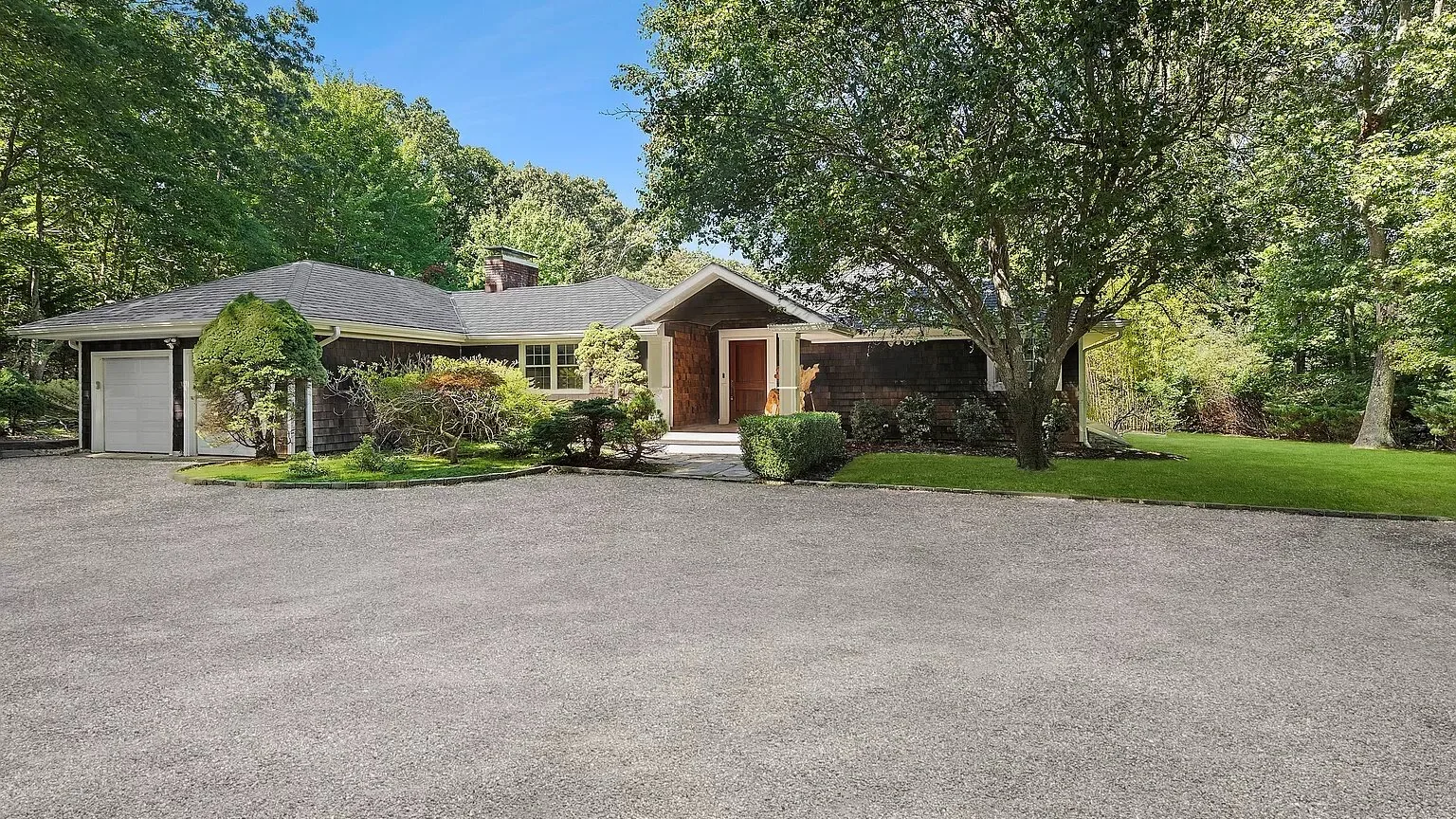 A single-story house with a paved driveway and leafy trees.