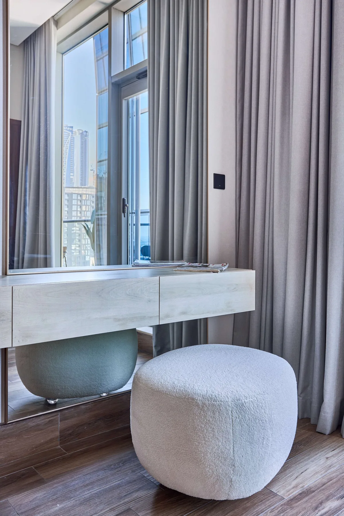 A window-side vanity and round upholstered stool sit beside floor-to-ceiling curtains.