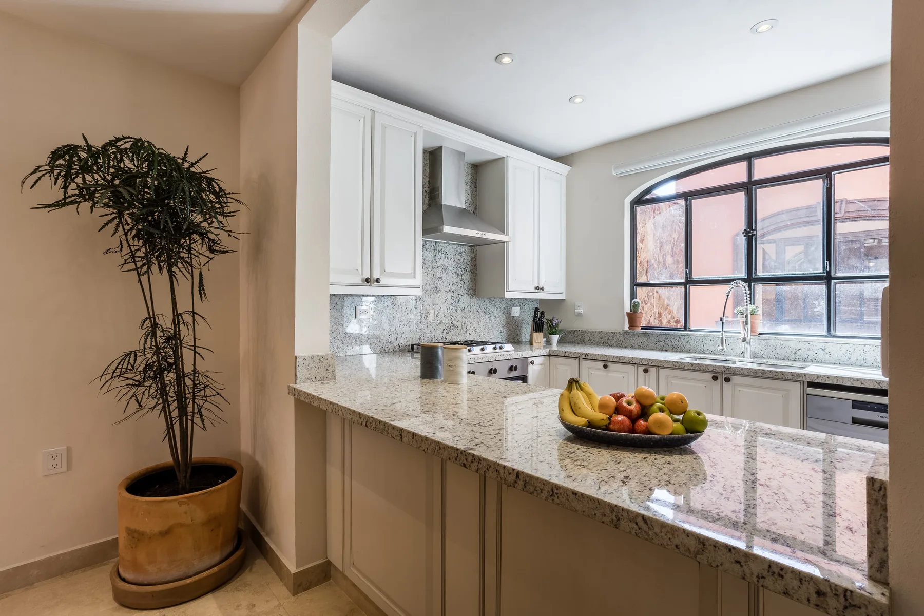 Una cocina luminosa con gabinetes blancos, encimeras de piedra y una gran ventana arqueada.