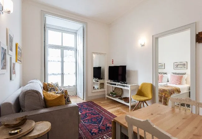 Una sala luminosa con asientos, un televisor y una puerta hacia un dormitorio.
