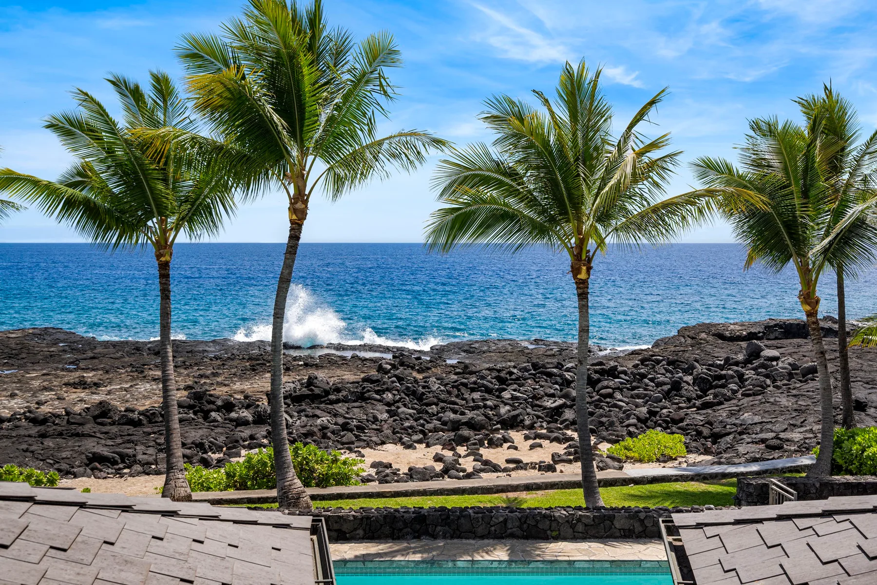 A tropical oceanfront scene with palm trees, dark rocky shore, and a swimming pool.