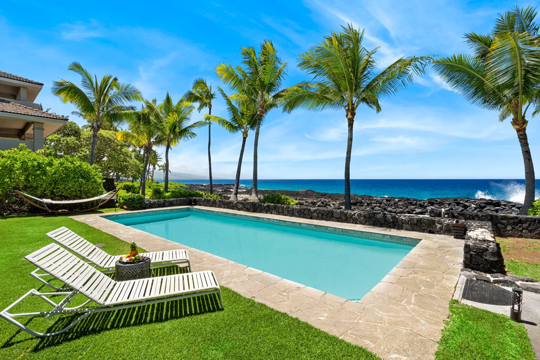 An outdoor swimming pool beside a grassy lawn, palm trees, and the ocean.