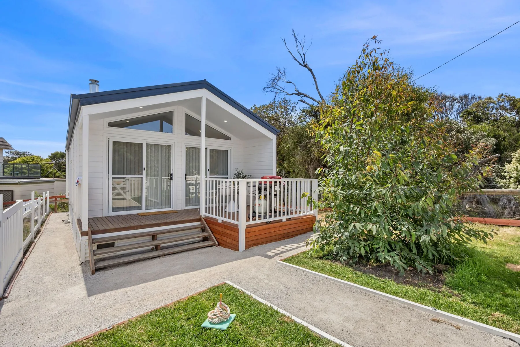 A small white house with a raised deck and steps sits beside a leafy garden.