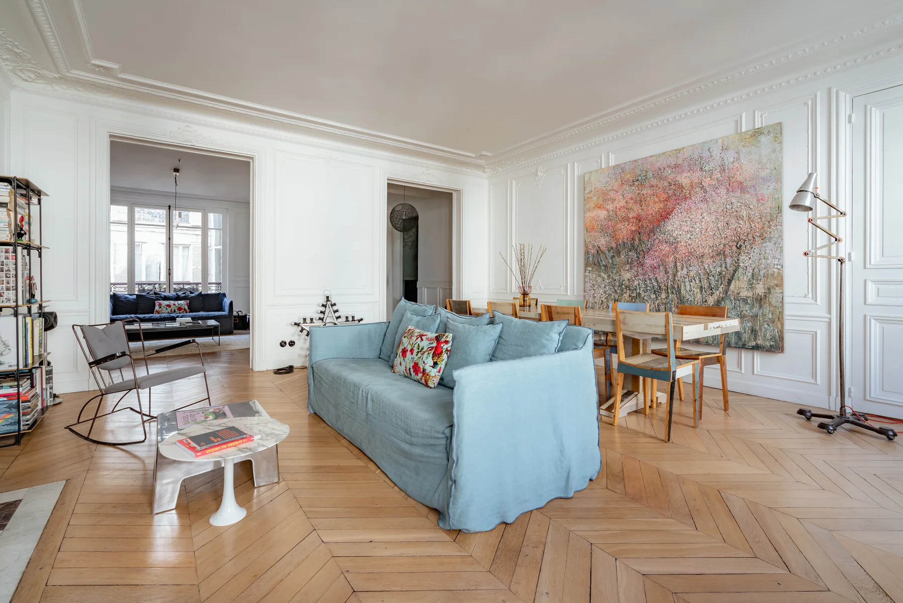 A bright living room has blue seating, a dining table, and a large colorful painting.