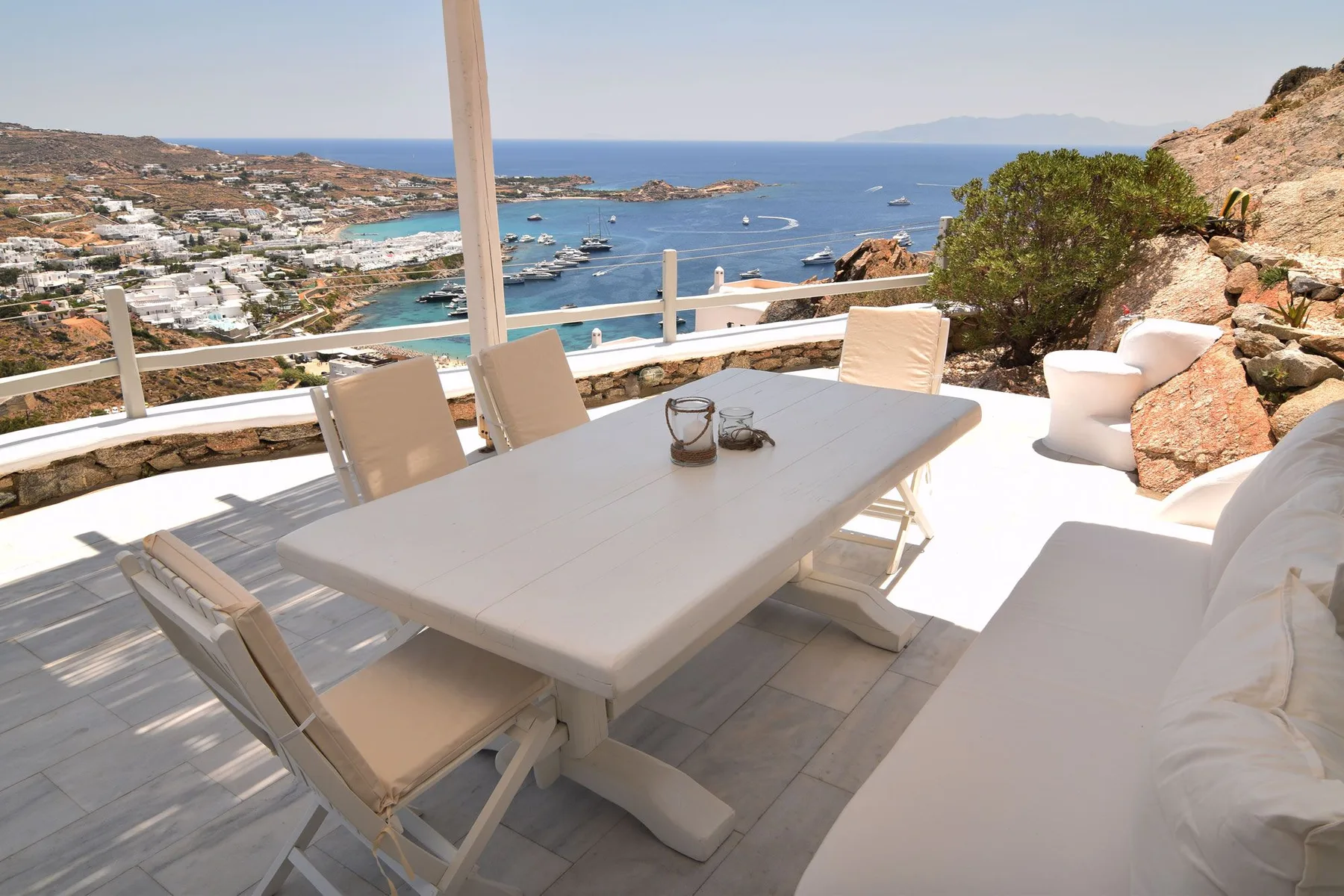 A sunny outdoor dining terrace overlooks a coastal bay, with a table and seating.