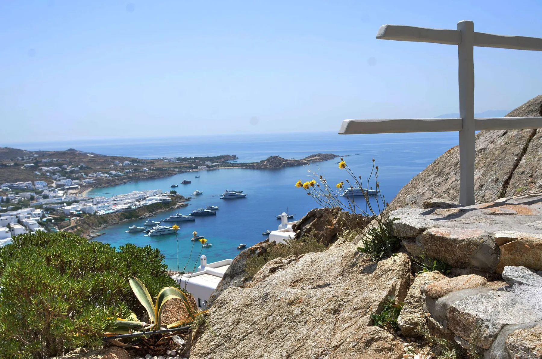 A coastal harbor with boats sits below a rocky hillside and wooden railing.