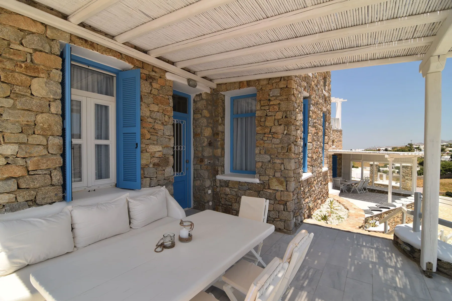 Covered stone terrace with white seating and blue shutters overlooking a sunlit landscape.