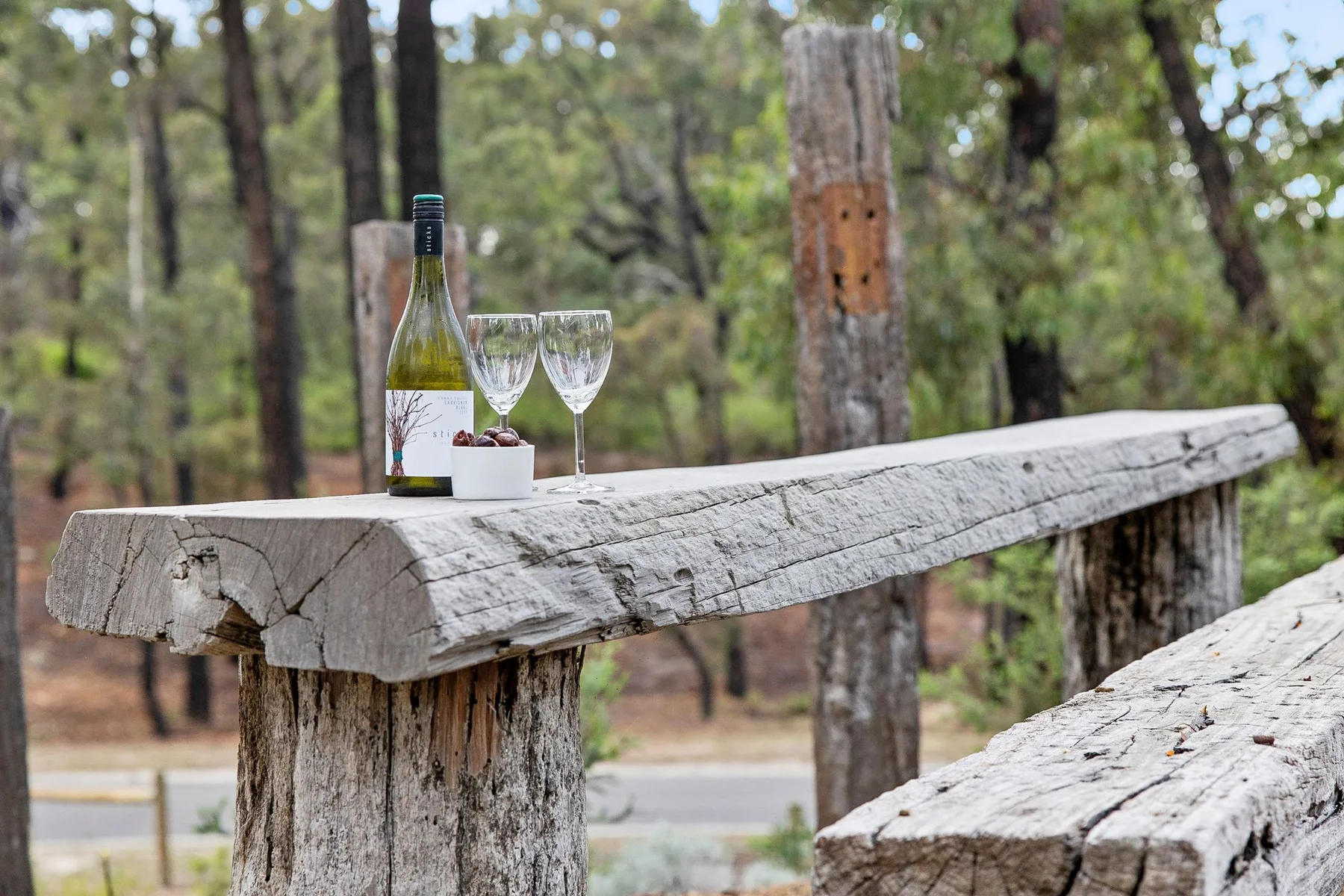 Una mesa rústica al aire libre sostiene una botella de vino, dos copas y un plato pequeño.