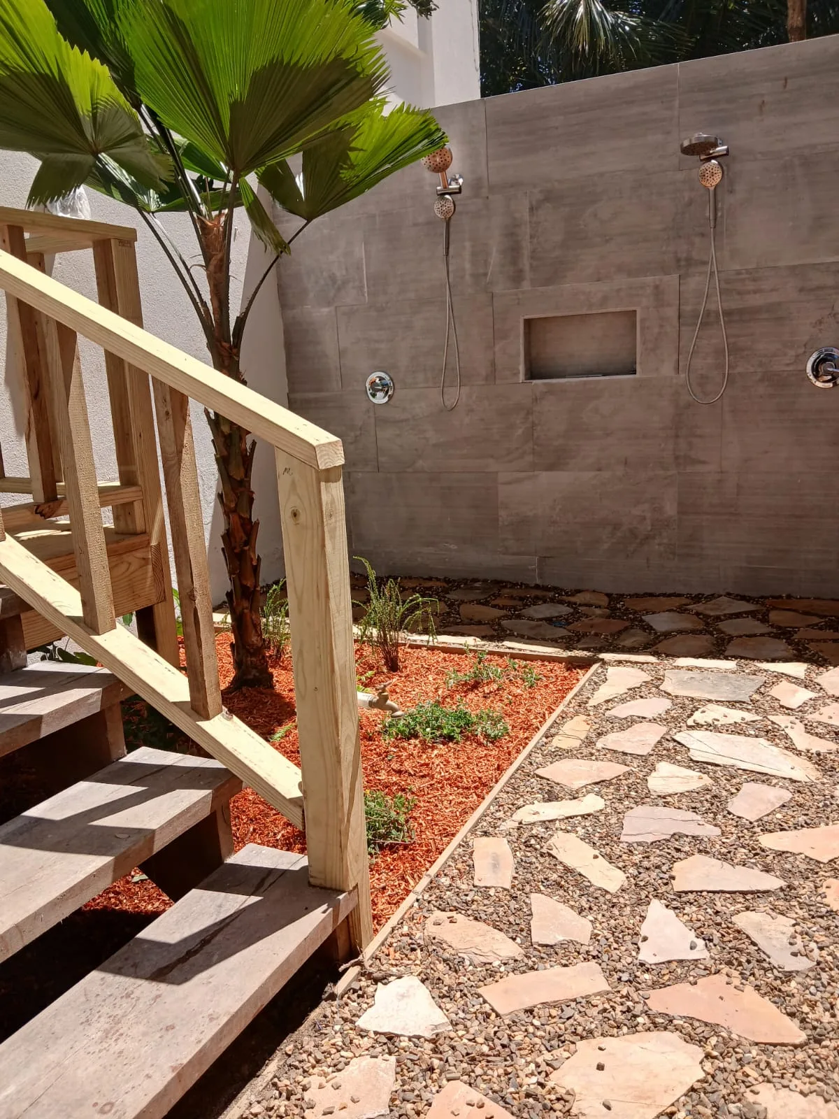 An outdoor shower area beside wooden steps, with a stone path and tropical plants.