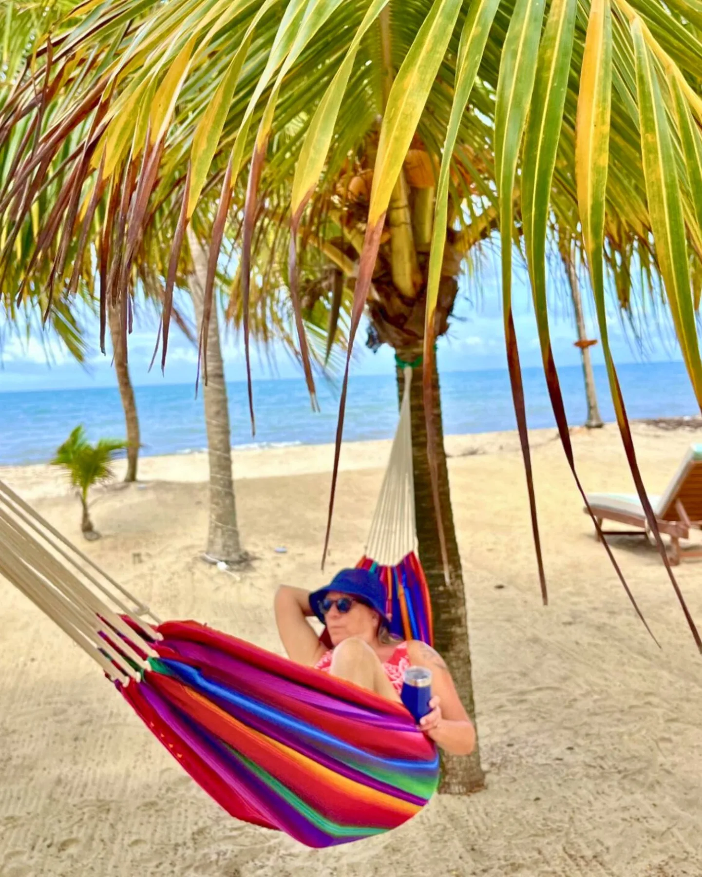 A person relaxes in a colorful hammock beneath palm trees on a sandy beach.