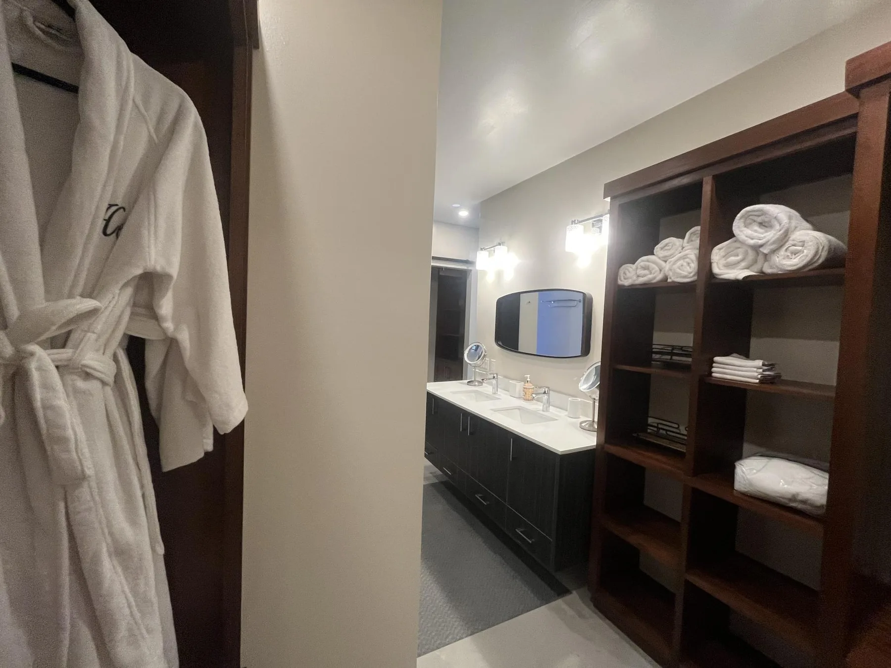 A bathroom vanity with a mirror stands beside open shelving stocked with folded towels.