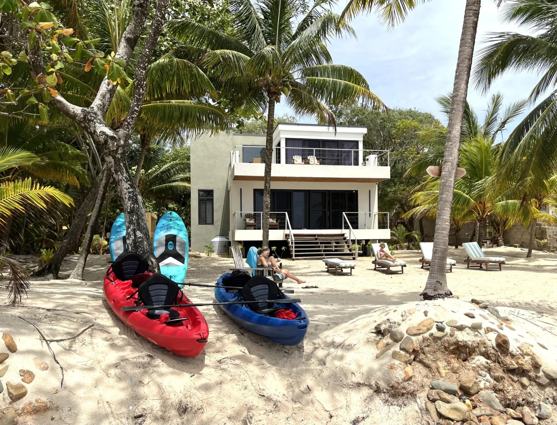 A modern beachfront house stands among palm trees, with kayaks on the sandy shore.