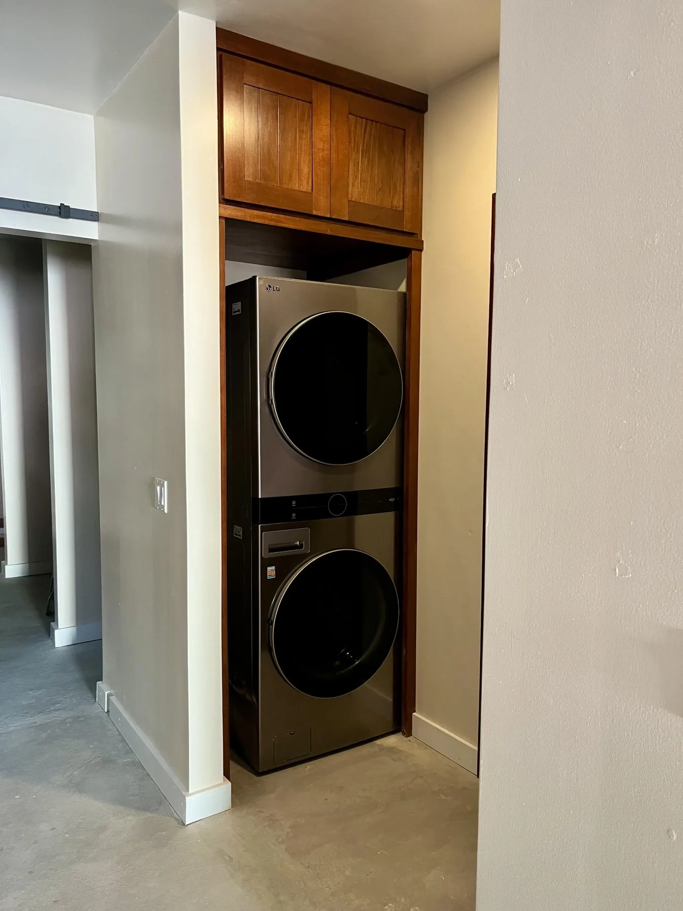 A stacked washer and dryer sit in a recessed laundry area beneath wooden cabinets.