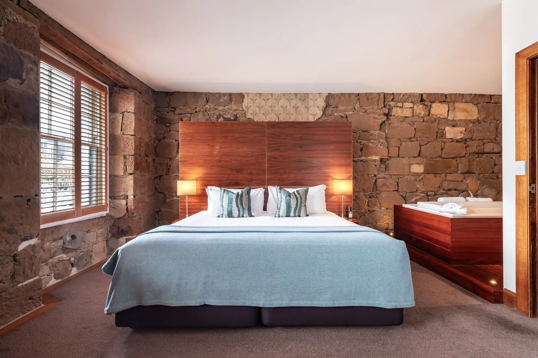 Representative hotel space: A bedroom with a bed against a stone wall and a window with blinds.