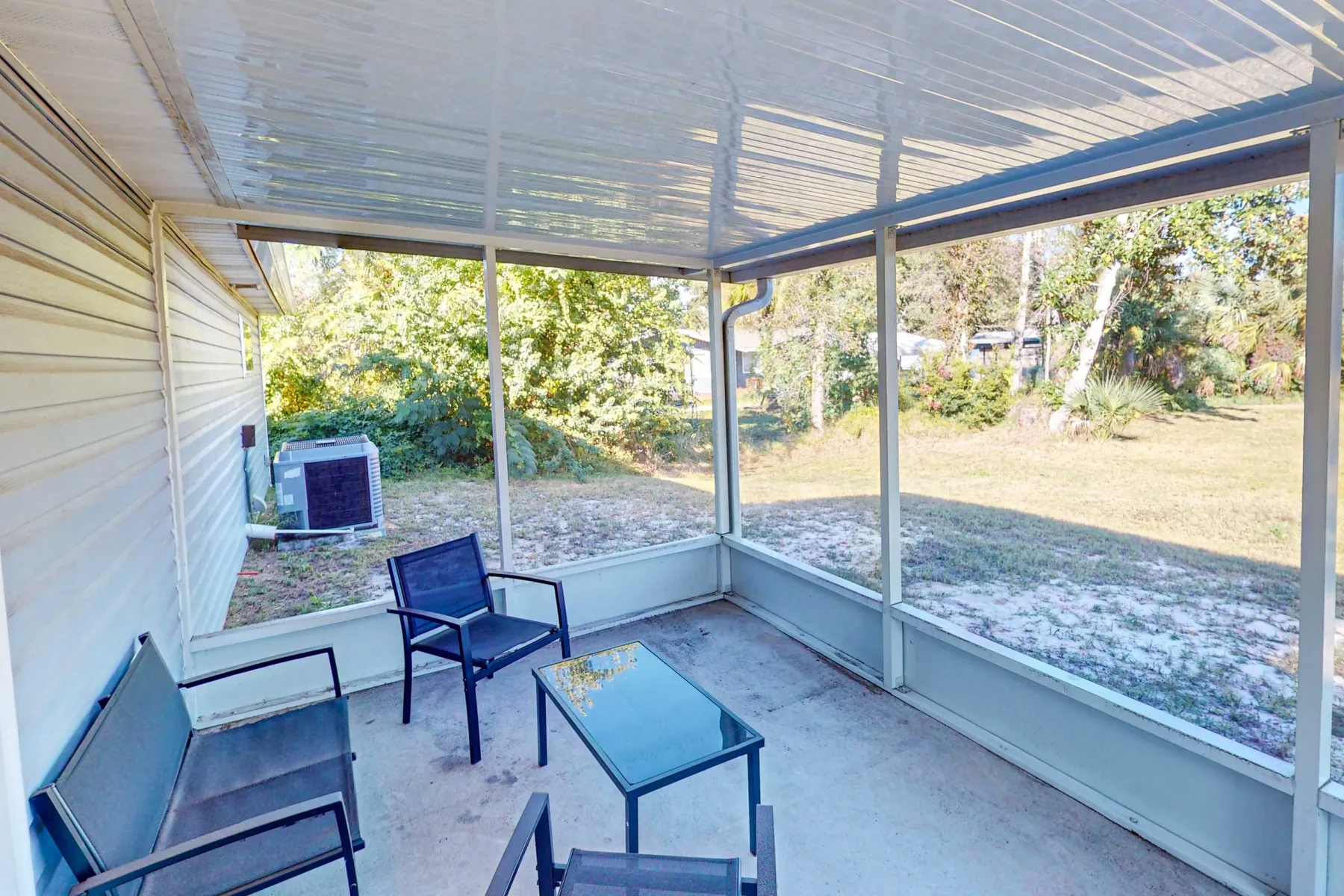 Un patio cubierto con asientos al aire libre y una mesa con tapa de vidrio da a un jardín con césped.