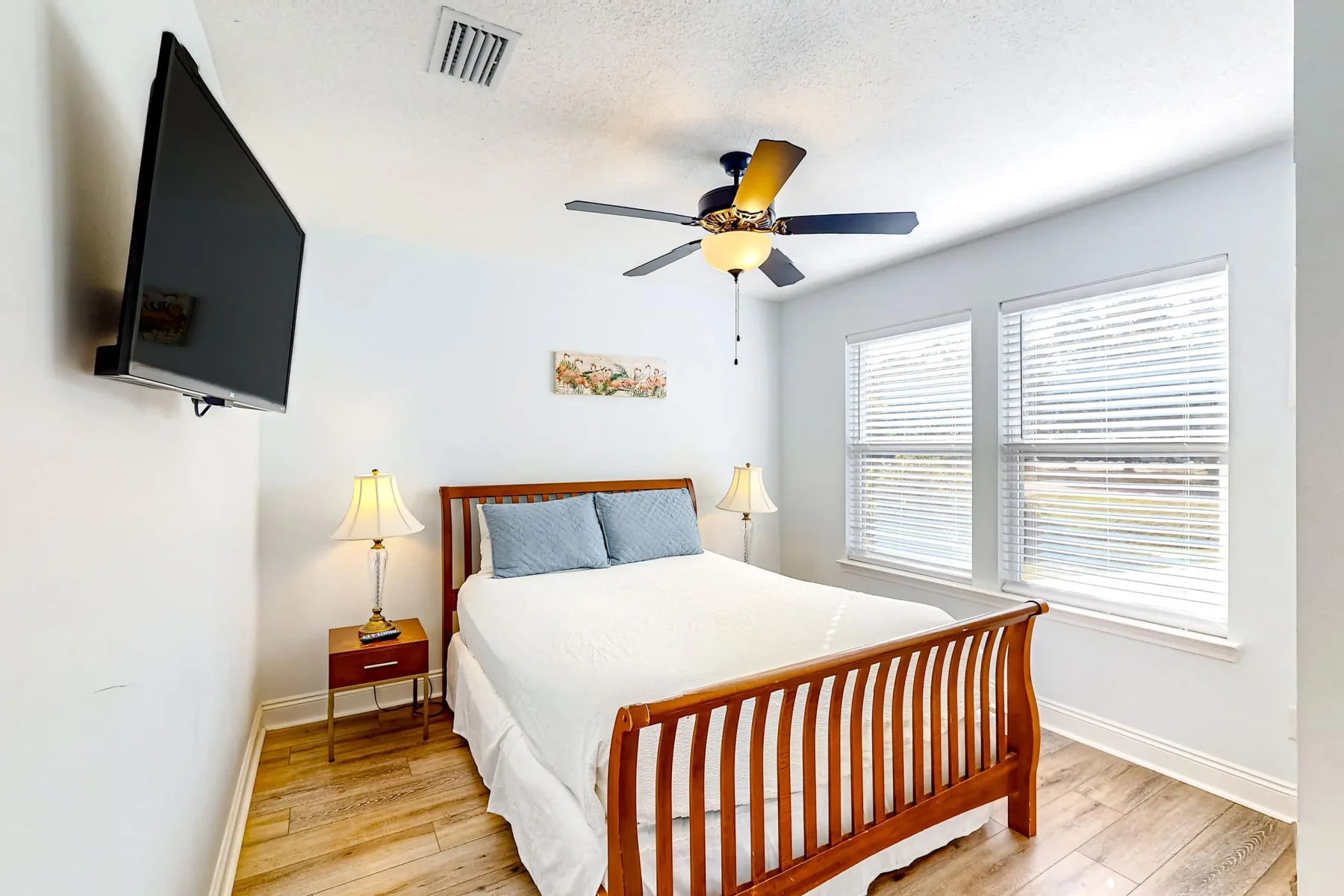 Un dormitorio luminoso con una cama de madera, lámparas de noche y dos ventanas grandes.