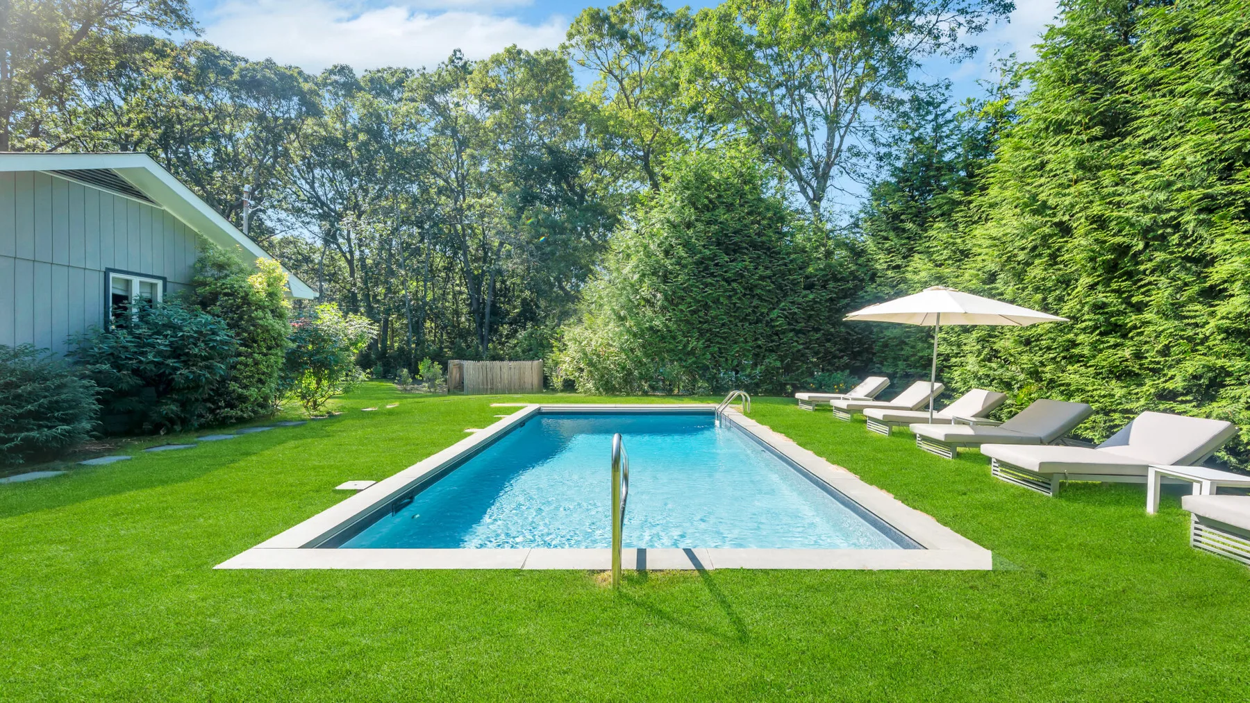 An outdoor swimming pool sits in a green yard beside lounge seating.