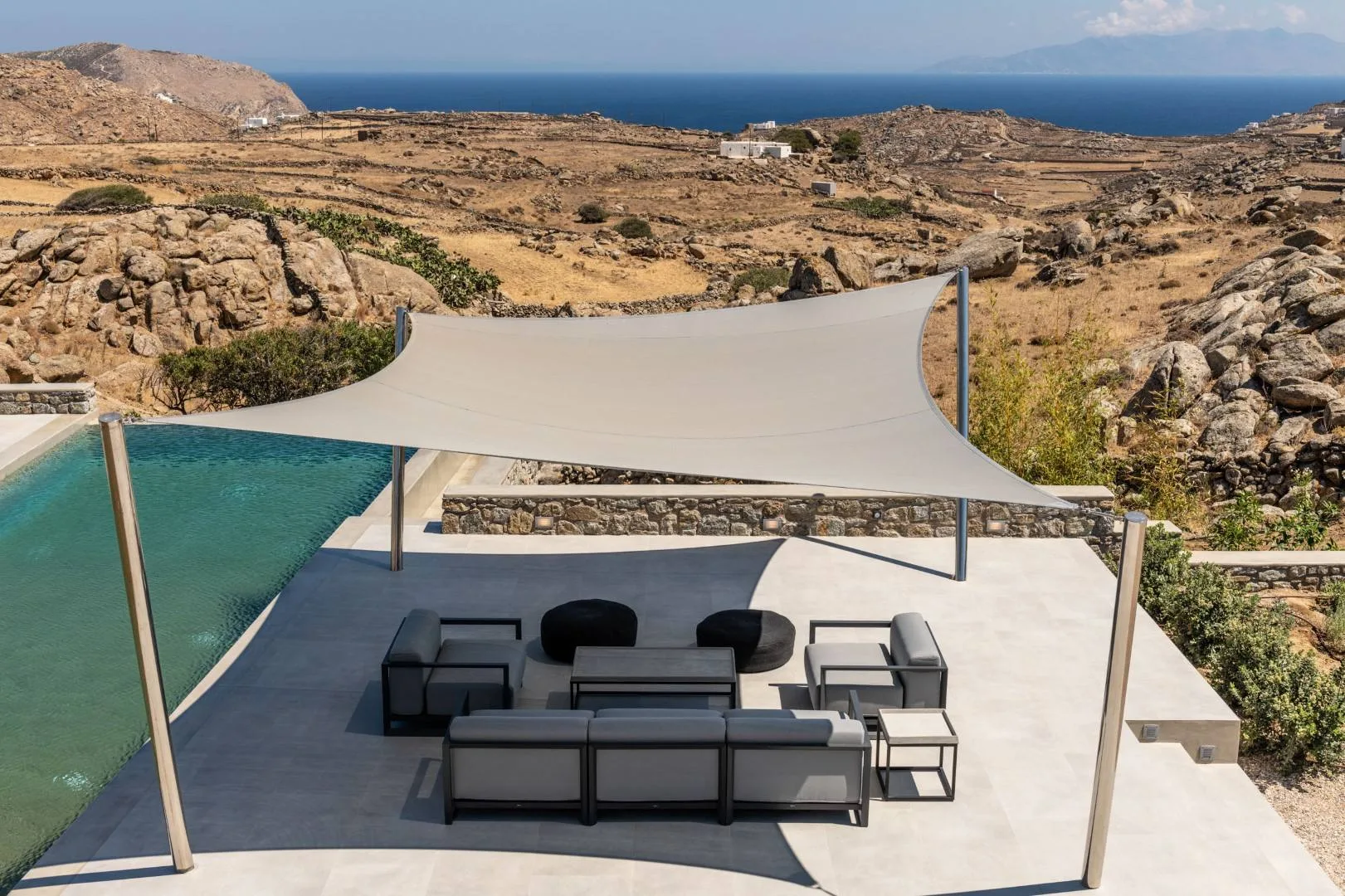 A shaded poolside patio with seating overlooks a dry, rocky landscape and the sea.