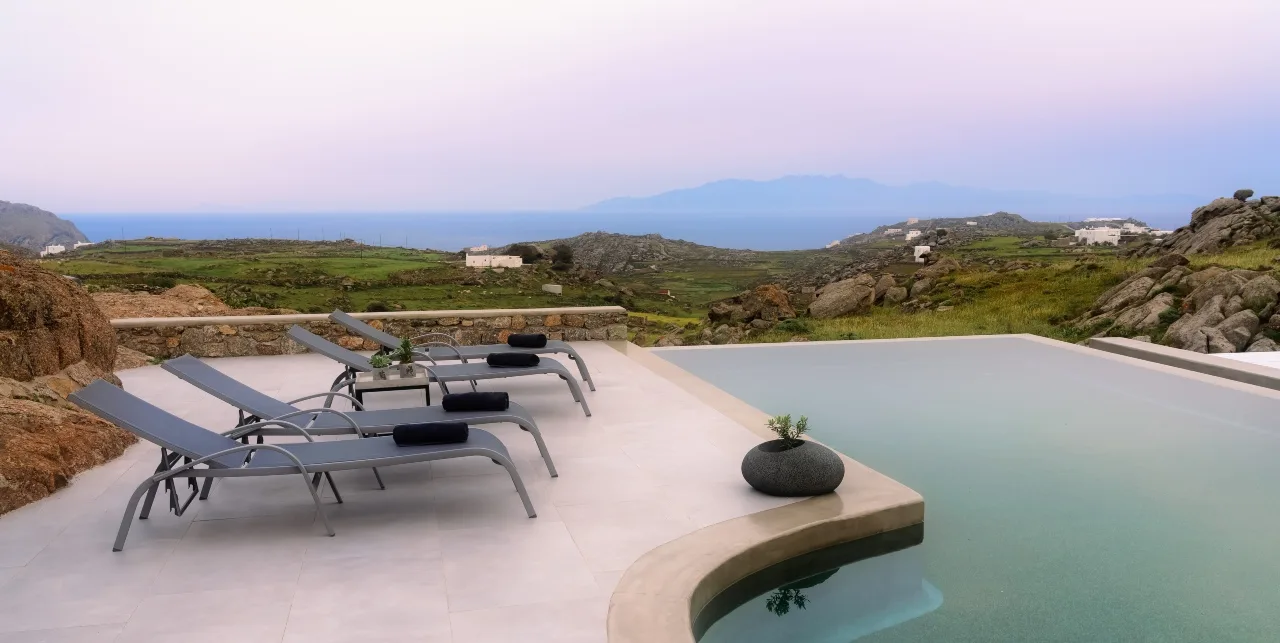A poolside terrace overlooks a green coastal landscape, with lounge seating beside the water.