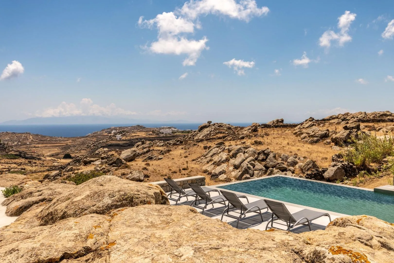 A swimming pool and lounge seating sit among rocky terrain overlooking the sea.
