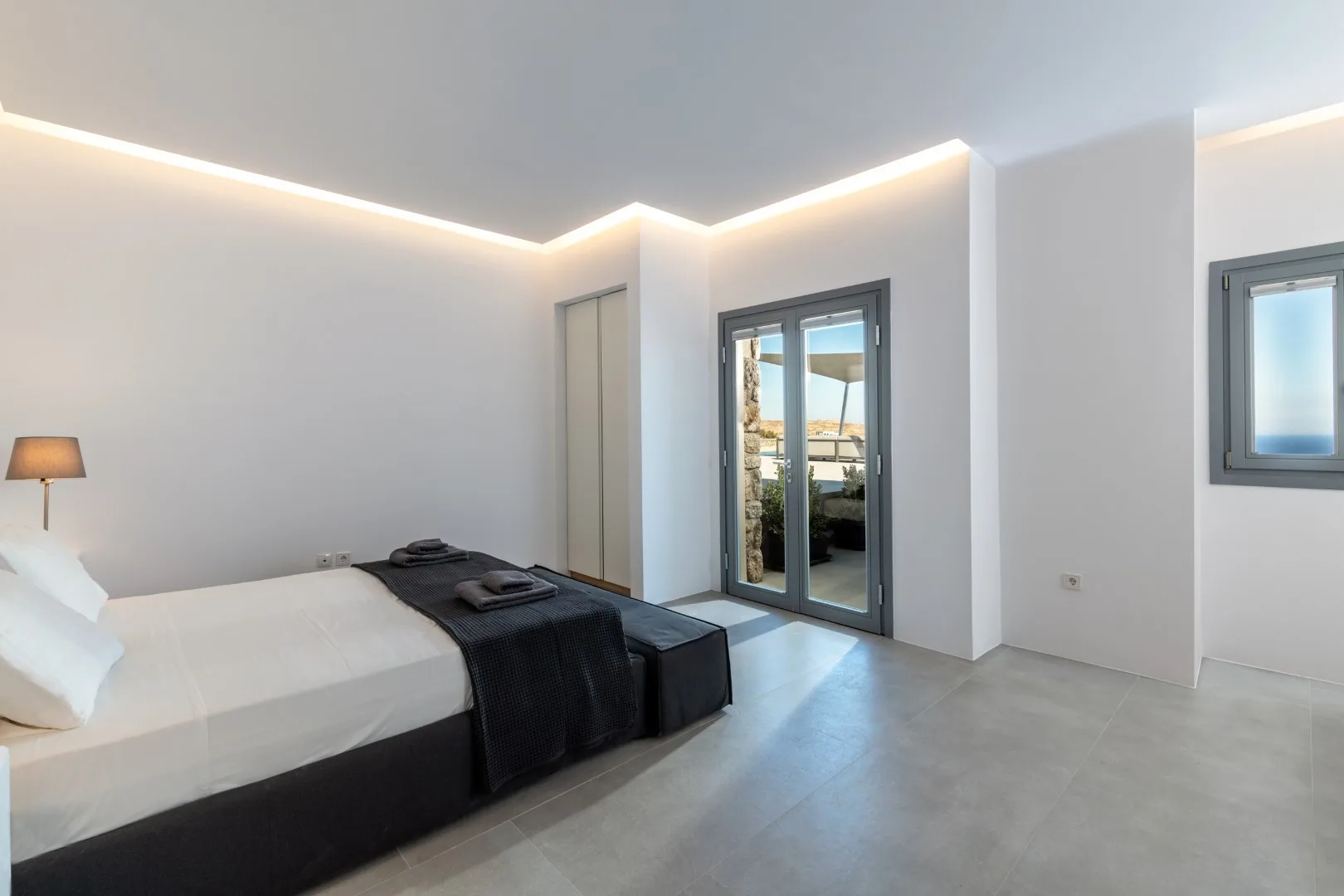 A modern bedroom with a bed, dark bedding, and glass doors opening onto a balcony.