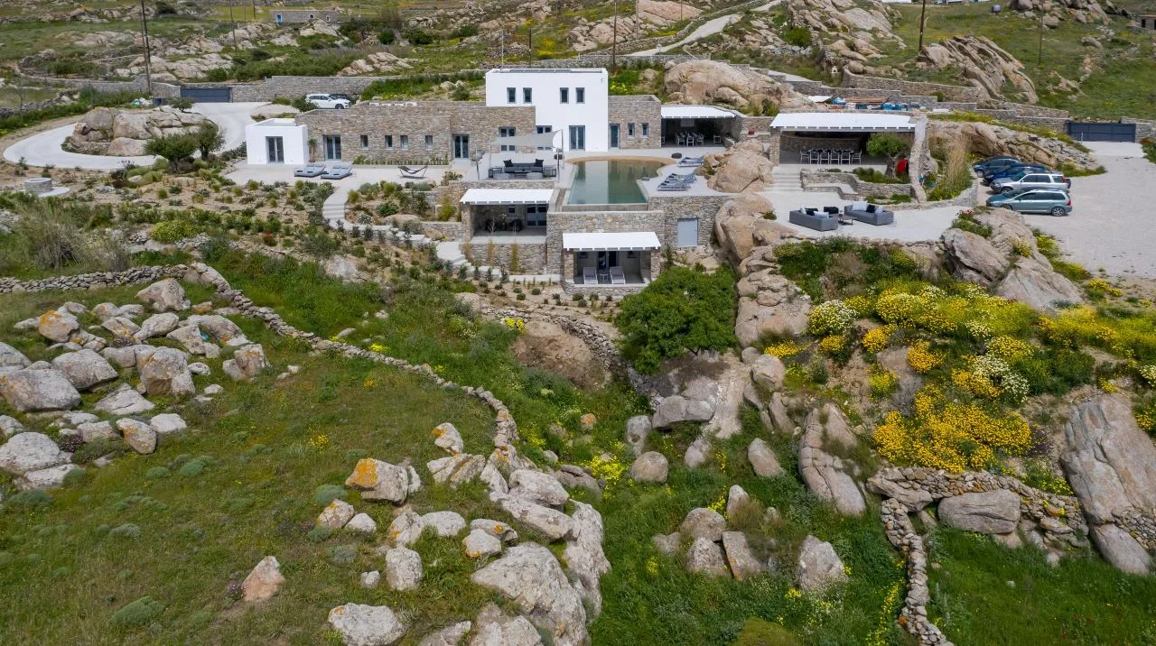 A modern stone-and-white building sits among rocky hills and green vegetation.