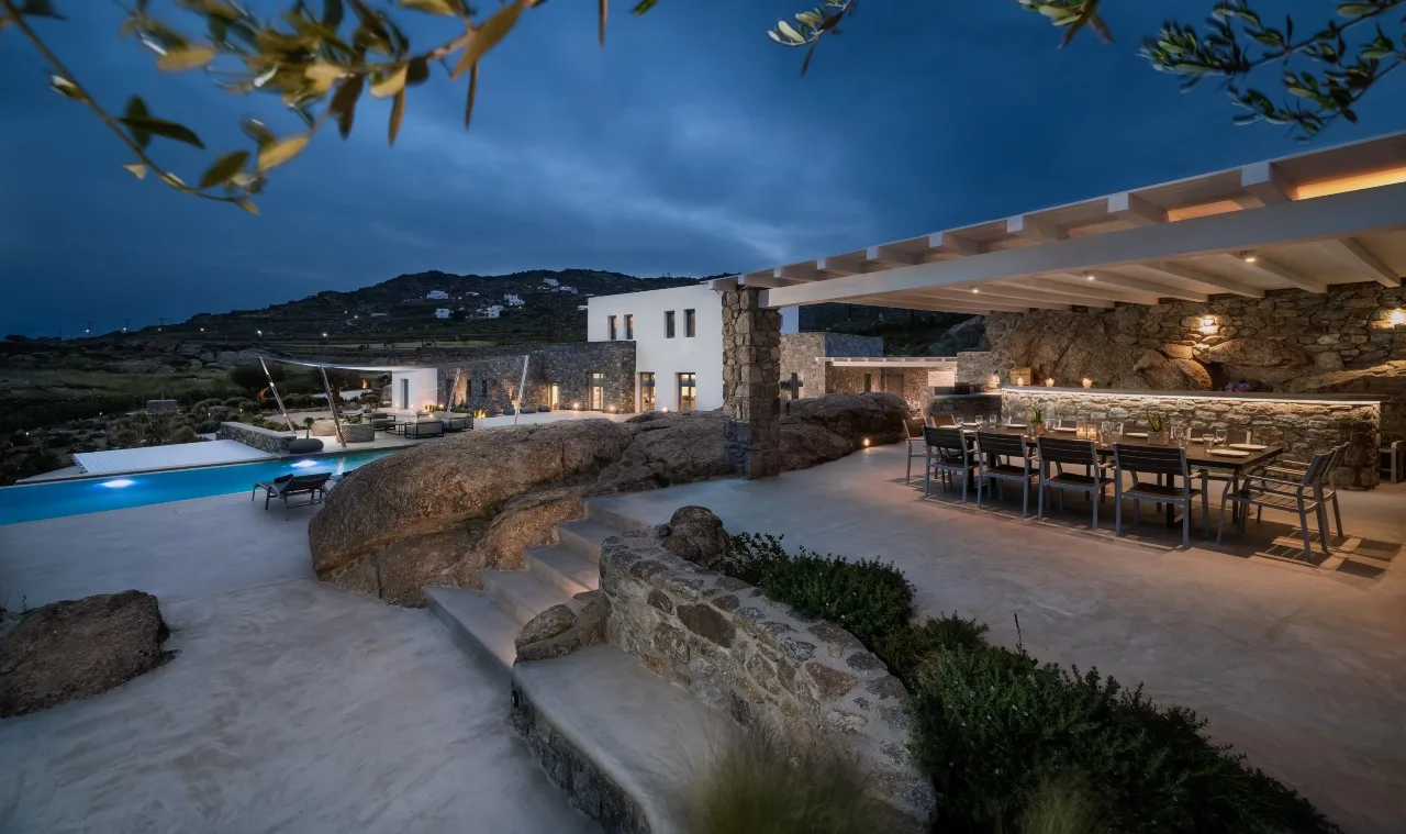 A modern outdoor patio with dining seating overlooks a pool and hillside at dusk.