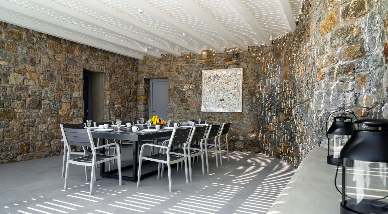 A stone-walled dining area with a long table and chairs beneath a covered ceiling.