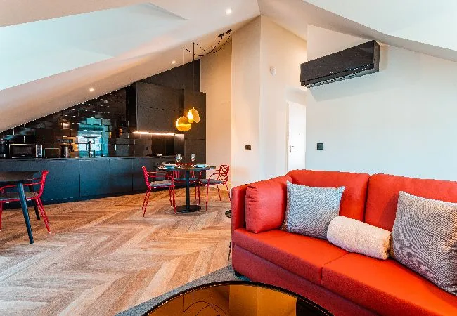 A modern living area with red seating beside a dining table and dark kitchen.