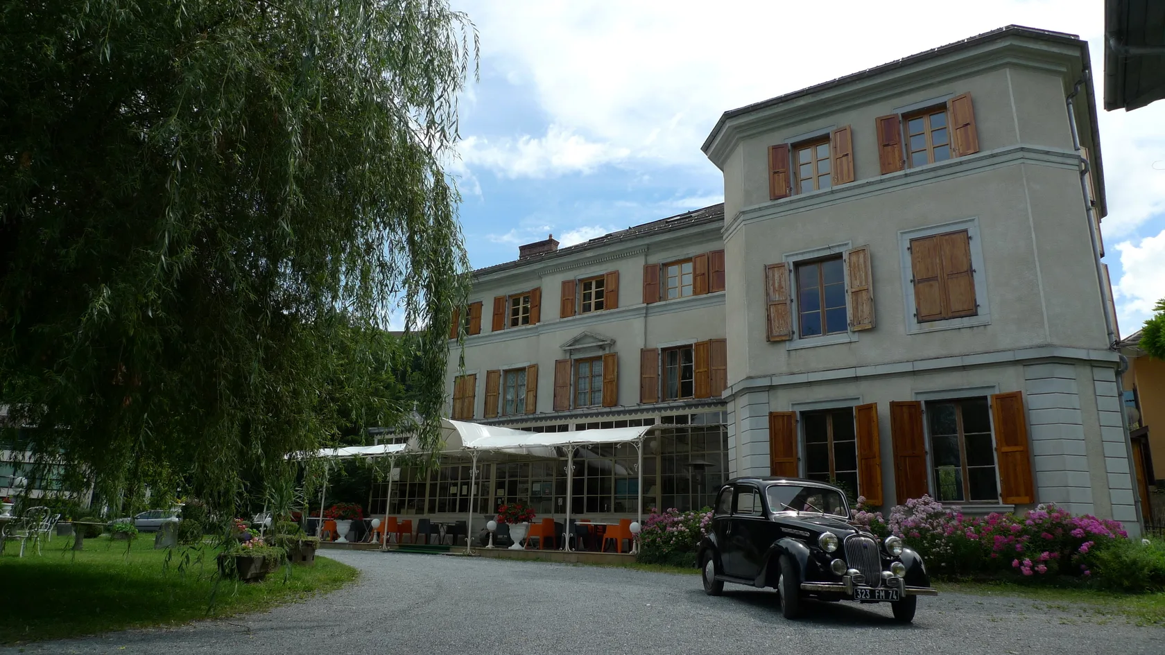 Espacio representativo del hotel: Un edificio grande con ventanas con contraventanas está junto a un jardín y mesas al aire libre.