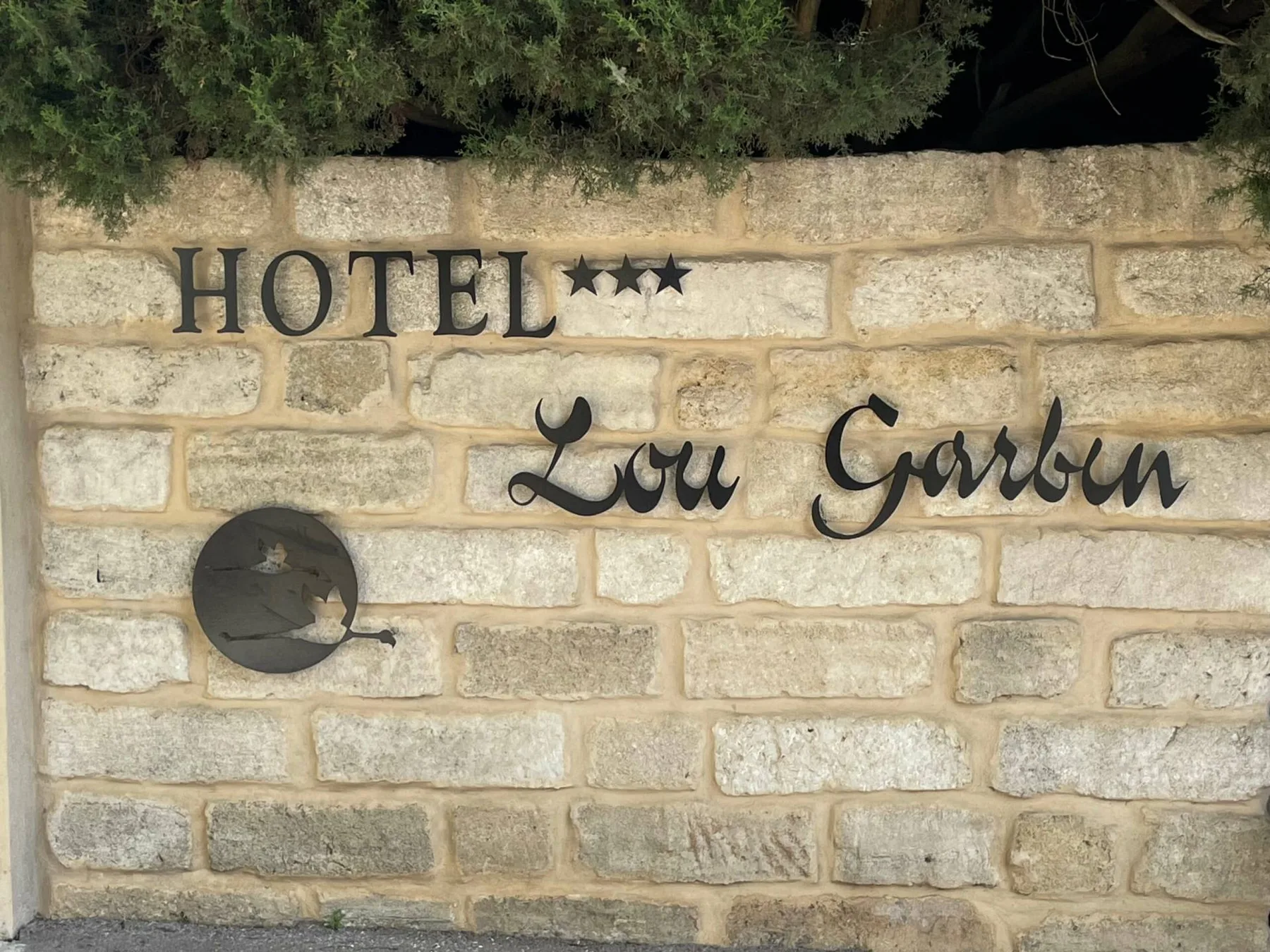 Representative hotel space: A stone hotel sign reads “Hotel Lou Garbèn” beneath overhanging tree branches.
