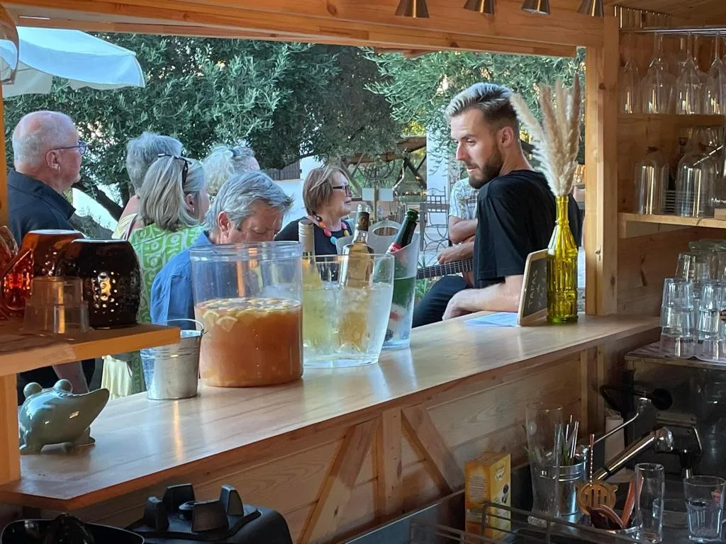 Representative hotel space: A wooden outdoor bar counter overlooks seated guests beneath leafy trees.