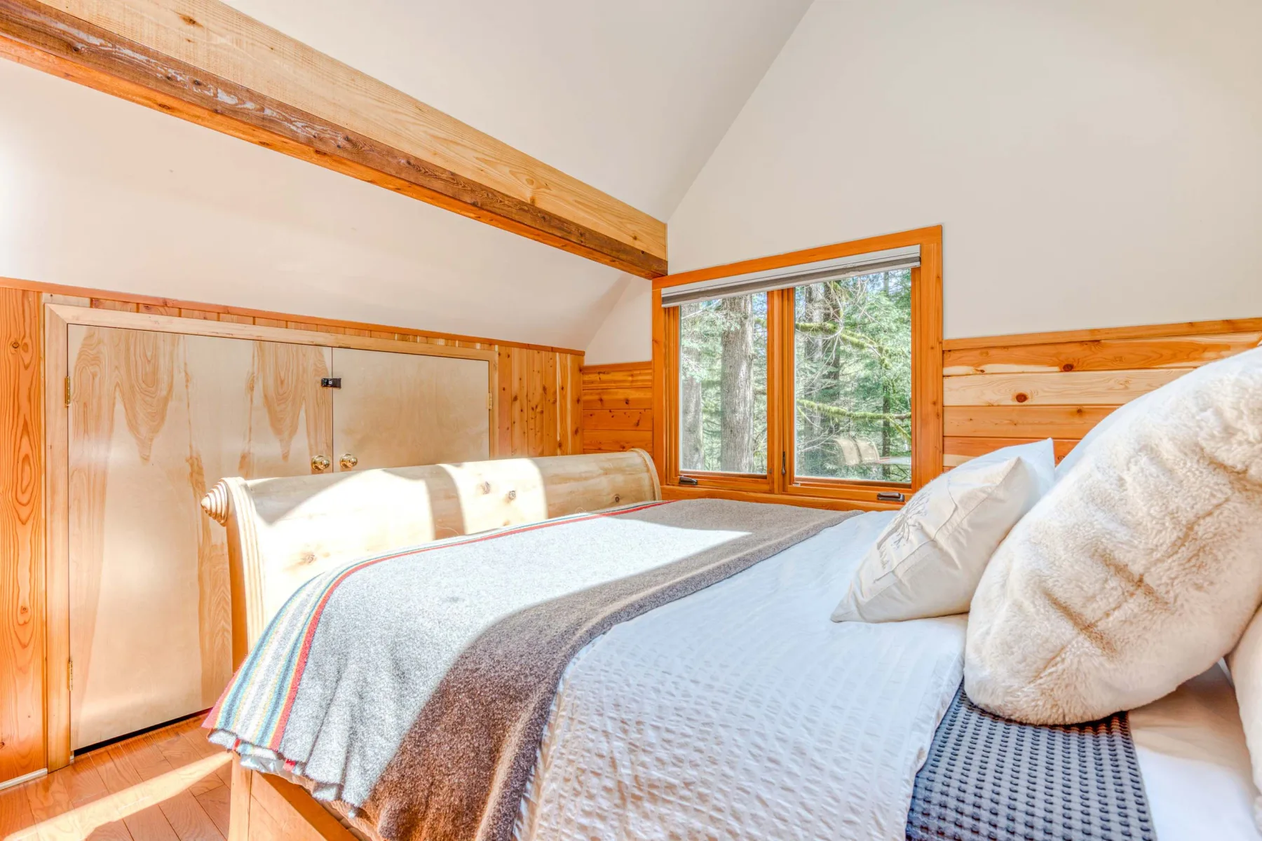 Un dormitorio con una cama bajo un techo inclinado y una ventana con vistas a los árboles.