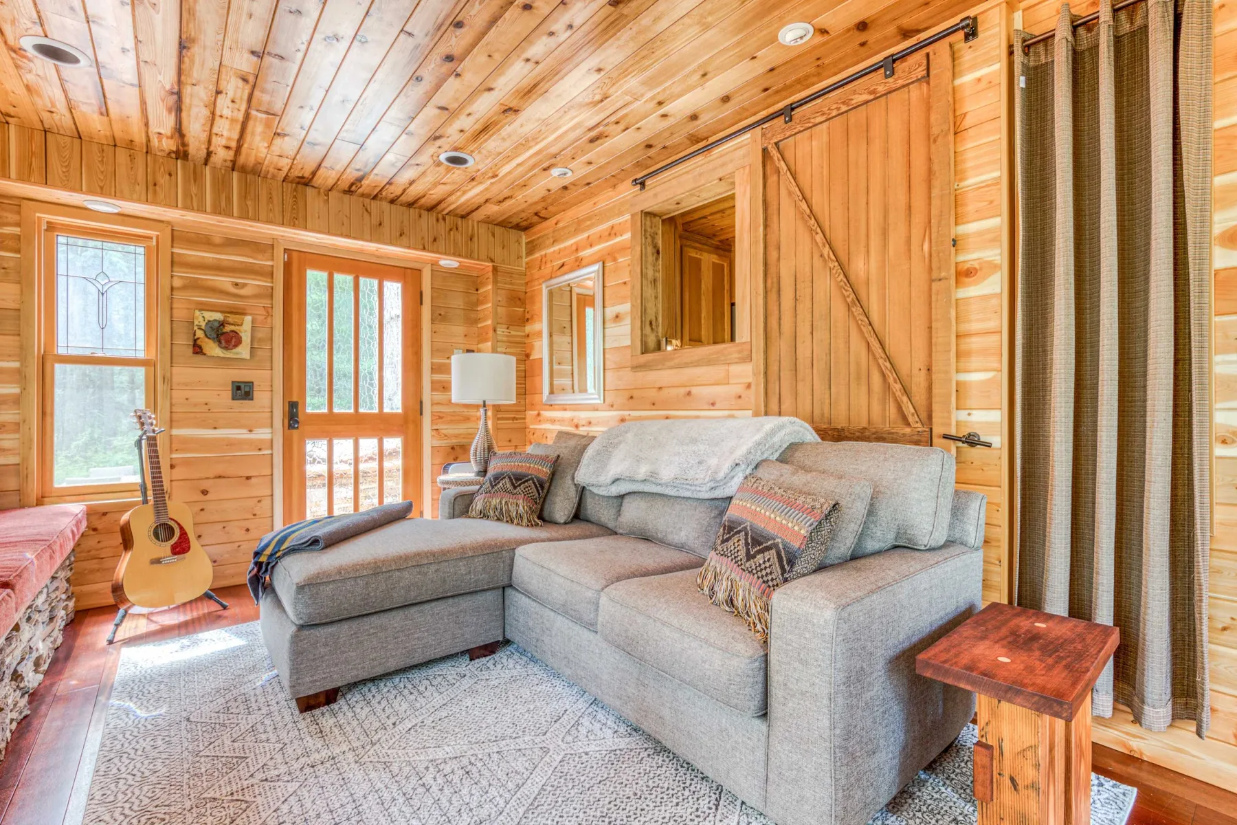 Una sala de estar revestida de madera, con asientos grises y una guitarra junto a la ventana.