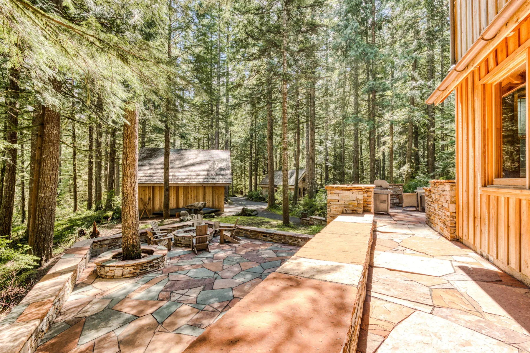 Un patio de piedra entre árboles altos, con asientos al aire libre y una cabaña de madera.