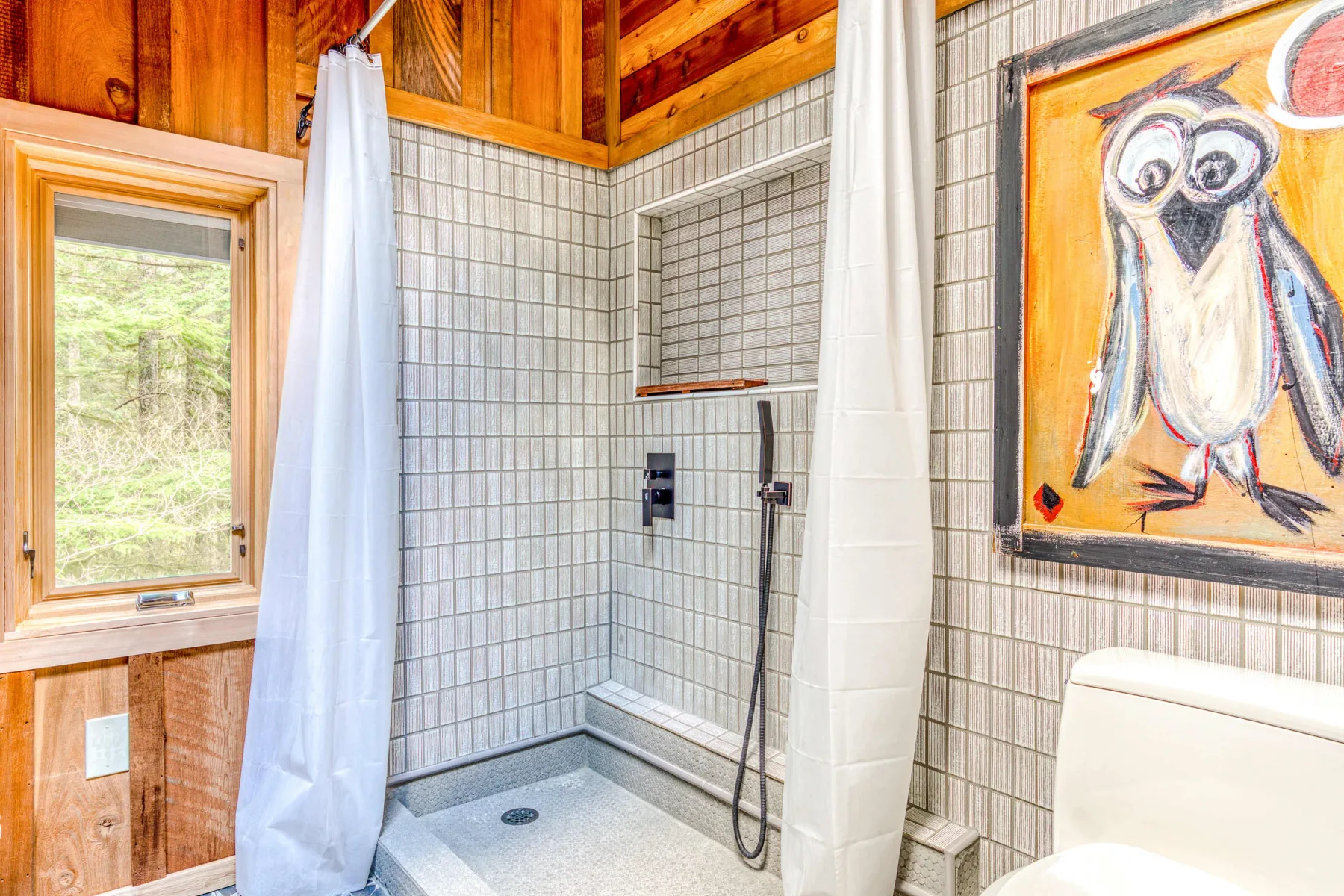 Zona de ducha de baño con azulejos, una ventana y una colorida obra de búhos.