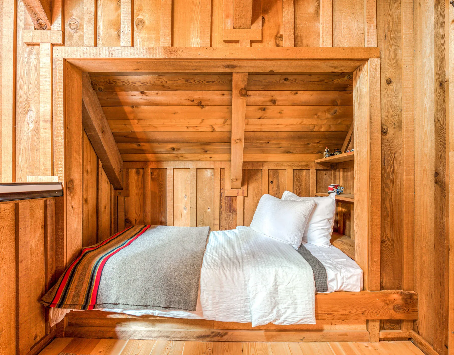 Un rincón de descanso de madera con una cama, ropa de cama blanca y una manta de rayas coloridas.